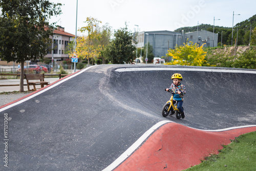 A young child rides a bike on a paved skate park ramp on a sunny day