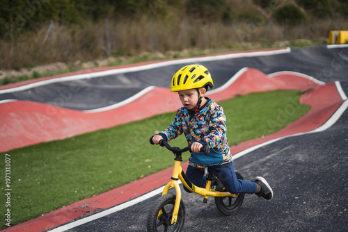 A young child riding a yellow balance bike on a winding paved path with green grass and red borders