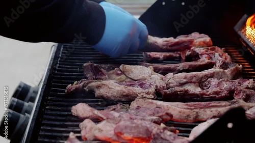 Professional chef carefully placing seasoned rosemary and garlic rubbed ribs on sizzling barbecue grill, methodically turning meat to achieve perfect even char and caramelization