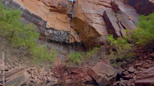 Footage captures the tranquil beauty of the Upper Emerald Pool in Zion National Park, Utah. The scene features calm, reflective water at the base of a massive, sheer sandstone cliff face. Lush green v