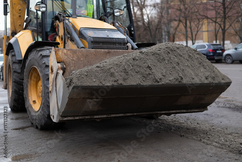Yellow excavator loader bucket filled with dark soil on a city street construction site.