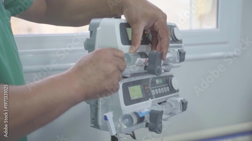 A side-profile, medium shot of a medical professional in green scrubs operating a dual-stacked syringe infusion pump. The professional's hands are shown precisely loading or adjusting a large syringe 