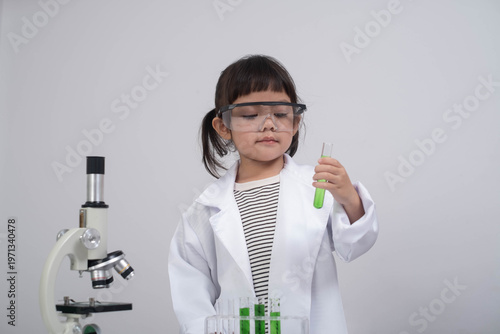 Little girl in lab coat and safety goggles holding test tube with green liquid beside microscope, STEM education concept, young scientist learning chemistry experiment isolated on white background.