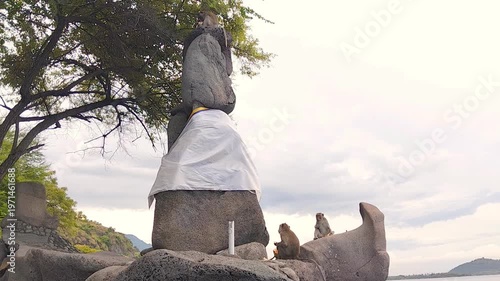 Ancient stacked stone monument, draped in sacred white cloth, observed by curious macaque monkeys on a serene tropical coast under a cloudy, overcast sky