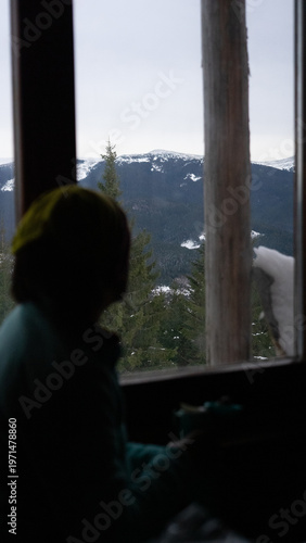 woman looking out the window at a beautiful landscape, snowy mountains and forest, winter