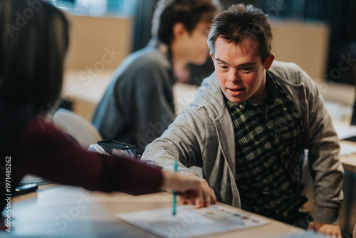 Young male student getting one-on-one help from a teacher at a desk. A teacher points to a worksheet while classmates work in the background.