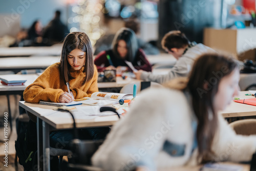 A young student studies intently at a desk surrounded by other students in a shared classroom. The scene shows a quiet group study environment with notebooks, pens and textbooks on tables.