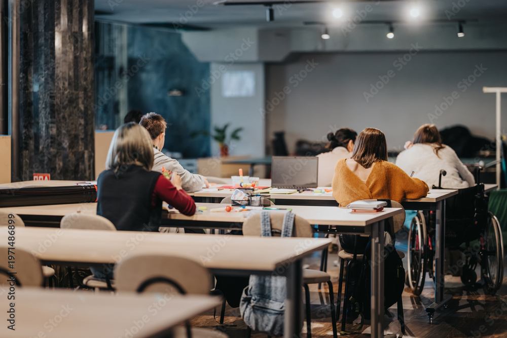 Obraz premium Adults study and collaborate at shared tables in a modern classroom or coworking workshop. A diverse group works with laptops, notebooks and art supplies while one person uses a wheelchair.