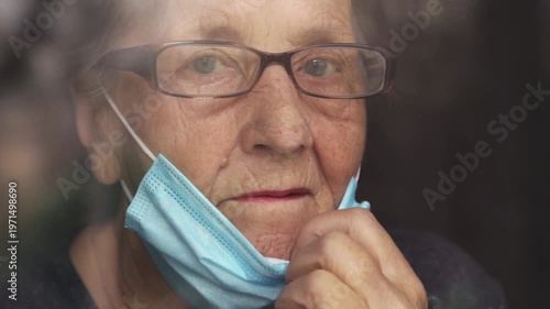 Portrait of an elderly woman in a window after quarantine with her mask removed. An old woman in a window with her mask down, after the pandemic.