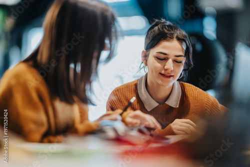 Two young women study together, sharing notes and focusing on their work. The pair collaborate and discuss ideas in a relaxed, indoor study session.