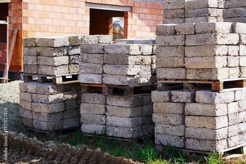 Construction blocks stacked on pallets, brick house in the background, construction site, muddy access road, scattered construction equipment and tools around.