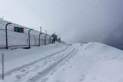 View of snow-covered ground with tire tracks leading towards a fence under a cloudy sky, Pir Chanasi, Azad Kashmir, Pakistan.