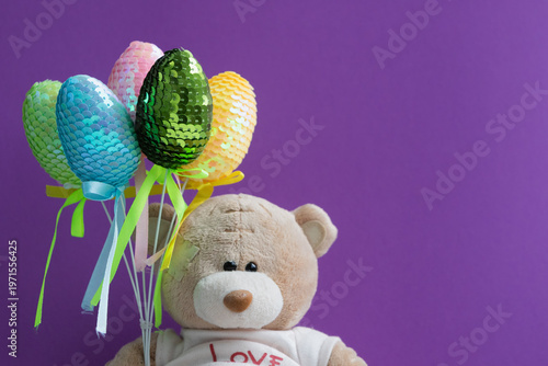 “A plush teddy bear with shiny colorful toy Easter eggs arranged in a bouquet on a lilac background. Easter holidays.”