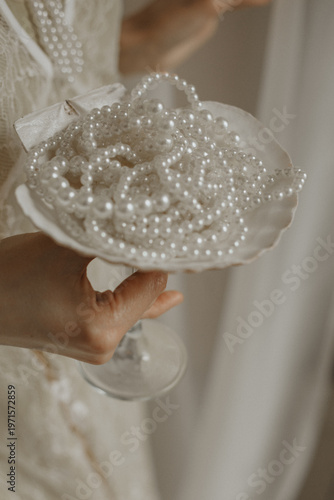 bride holding a collection of pearl necklaces in a vintage glass dish. Soft natural light and warm tones create an elegant and timeless wedding atmosphere, focusing on accessories and luxury details