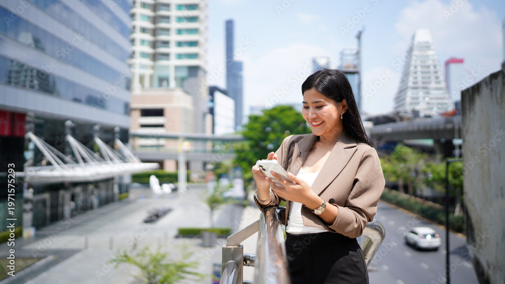 Fototapeta premium Successful Asian businesswoman smiling while using smartphone standing on balcony surrounded by modern skyscrapers in the urban financial district