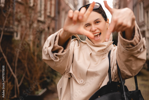 Excited young woman in stylish beige trench coat framing view with her hands and winking at camera, cheerful female traveler exploring European city street, professional photography of happy lifestyle