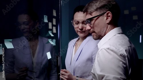 Business professionals brainstorming new ideas and strategy, collaborating on project development using sticky notes on a glass board in a dark modern office meeting room