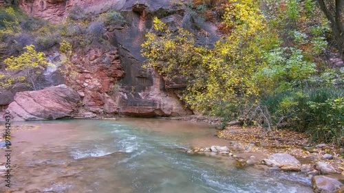 Footage captures the serene beauty of the paved Riverside Walk trail in Zion National Park, Utah, during the vibrant autumn season. The path follows the Virgin River as it winds through the narrow can