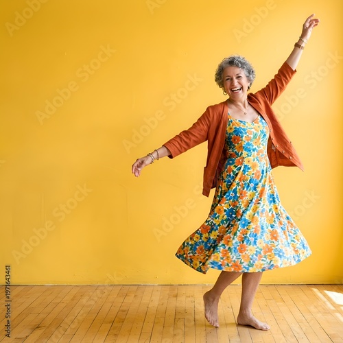 Joyful senior woman in floral dress and orange cardigan dancing barefoot on wooden floor with vibrant yellow background