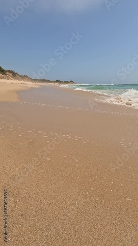 A low-angle vertical view of a pristine coastline with turquoise waves crashing on golden sand, dunes, and blue sky. The wild and famous beach is called Bolonia, in Cadiz, Andalusia, Spain