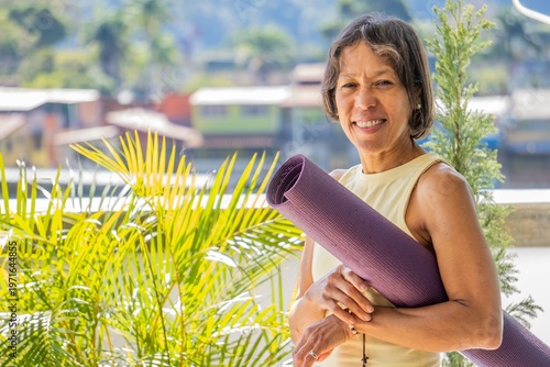 Smiling mature woman holding a purple yoga mat outdoors, concept of active aging, senior wellness, and healthy lifestyle. A portrait of a happy mature woman with short dark hair, wearing a yellow tank