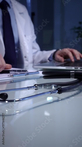 Stethoscope laying on a white reflective desk while doctor man working in clinic office. Typing on laptop and writing notes. Medicine concept