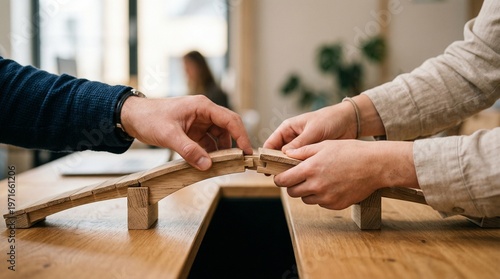 Wooden bridge model pieces being joined by two hands across a desk gap in warm natural light