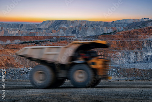 Fotografía Dump truck speeding past massive open pit mine walls at dusk, gold mining indust