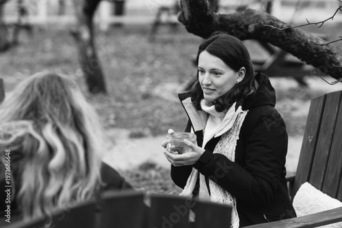 Two pretty caucasian women in their 40s wearing warm clothes spending time outdoors in autumn park cafe. Mature females enjoying personal time, talking, drinking tea, relaxing and breathing fresh air.