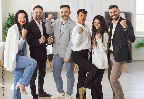 Εκτύπωση καμβά Group of young multiethnic teammates stands proudly in office, looking at camera and raised fists in triumphant Yes gesture, celebrate shared moment of success, achieving joint goal