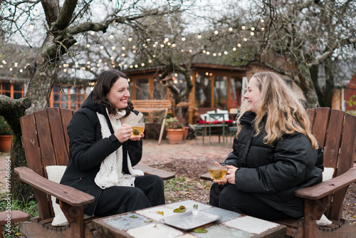 Two pretty caucasian women in their 40s wearing warm clothes spending time outdoors in autumn park cafe. Mature females enjoying personal time, talking, drinking tea, relaxing and breathing fresh air.
