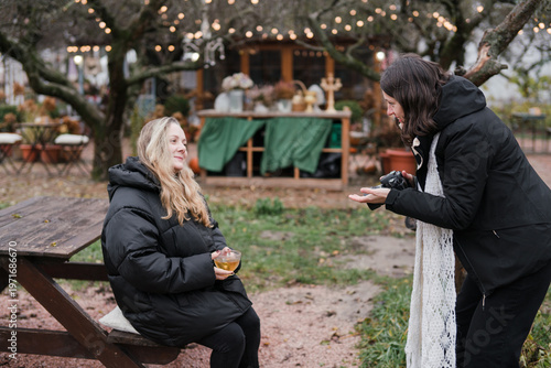 Caucasian woman photographer in her 40s wearing warm clothes taking photos of her friend in autumn park. Relaxed mature females spending time outdoors, enjoying leisure time, walking and posing.
