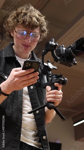 Vertical screen Videographer studies script on smartphone holding camera on gimbal looking confused struggling to understand how to execute idea planning scene in studio
