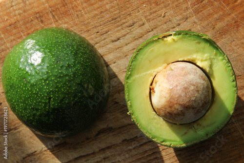 Quadro su tela Fresh halved avocado on a rustic wooden cutting board, displaying vibrant green flesh, a large seed, and bumpy skin under natural sunlight