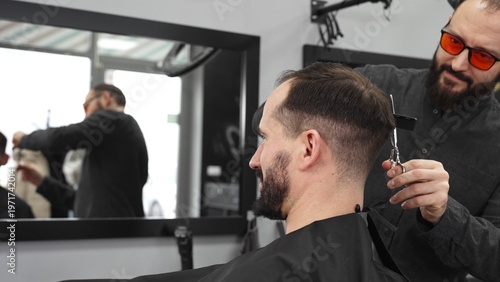 Fotografie Skilled barber working on male client's hairstyle, carefully combing and prepari