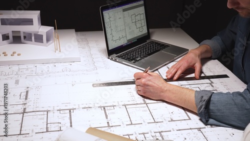 Man engineer working on construction development, drawing architectural blueprints and using a laptop with design software, surrounded by hard hats and a building model in a darkly lit studio, close
