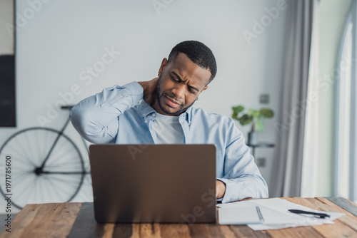 Young Black man rubs his neck while working on a laptop in a bright home office, showing frustration and physical discomfort from prolonged computer use and poor posture.