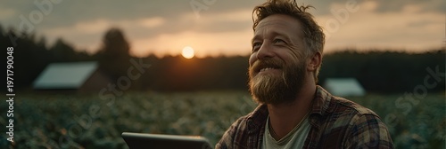 Bearded farmer with tablet smiles at the sunset over the field and barns, envisioning a bountiful harvest and successful agricultural business.