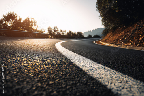 Low angle view of winding asphalt road with white lane marking illuminated by warm sunset light, detailed textured surface and dramatic curve through countryside landscape
