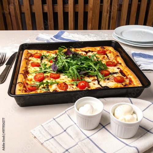 Square pizza with tomatoes and arugula, sides of cheese. Table setting