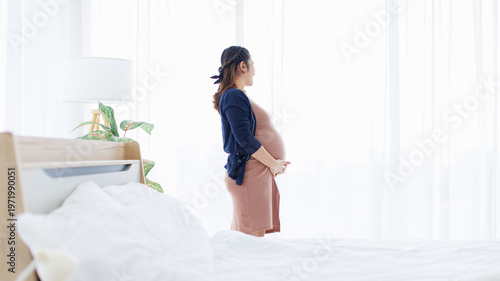 Asian young pregnant woman standing near window in living room at home smile pose touching belly care peaceful comfortable love healthy happy pregnancy motherhood maternity tummy wellness mother day.