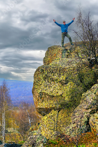 an elderly happy tourist stands on a high rock against the backdrop of the Ural mountains on a summer day