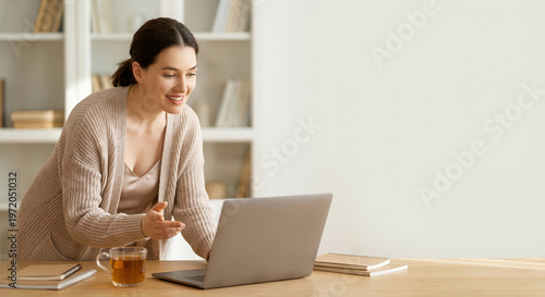 A woman is working on her laptop at a desk, with a cup of tea and notebooks nearby. 