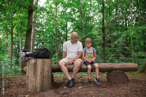 Father and little son resting on wooden bench in green forest during family outdoor walk