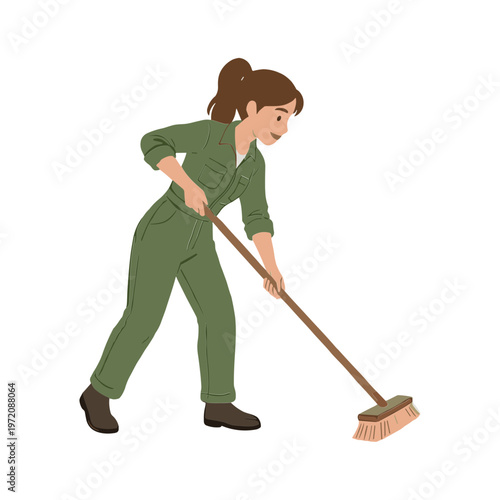A woman wearing green overalls sweeps the floor with a broom indoors.