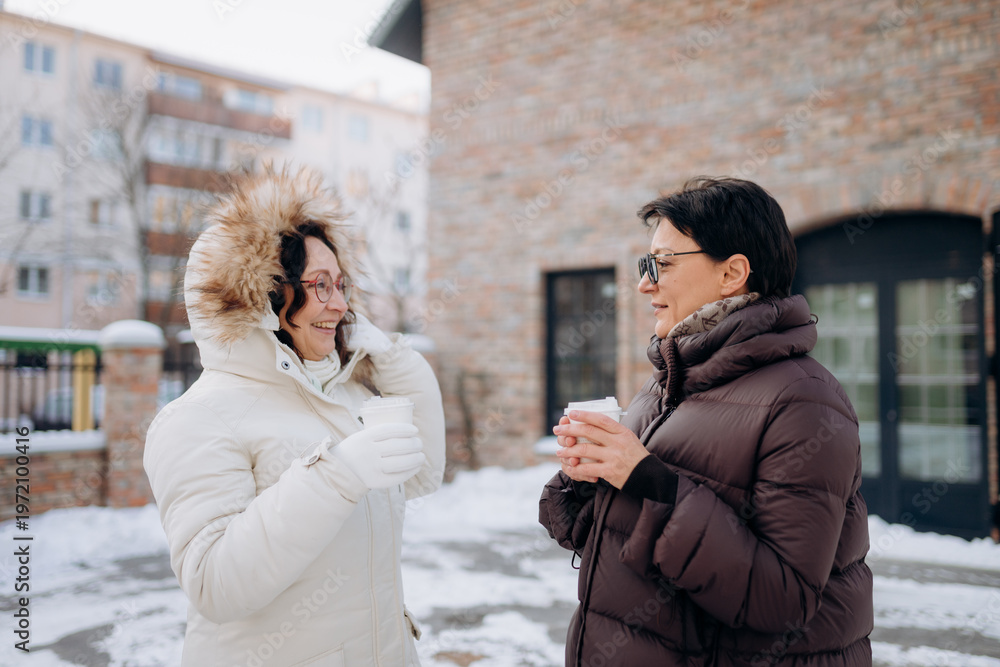Fototapeta premium Two friends stand outside in a winter wonderland, wrapped in cozy coats. They share smiles and warm drinks while enjoying a chilly yet cheerful day together in the city.