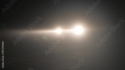 Car headlights cutting through dense fog at night, bright vehicle beam piercing thick mist on dark road creating dramatic low visibility driving atmosphere