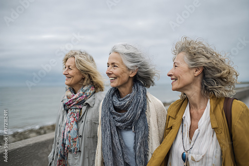 Three mature retired women strolling a seaside promenade with wind - swept grey hair, colorful scarves and a mustard jacket, smiling toward the ocean