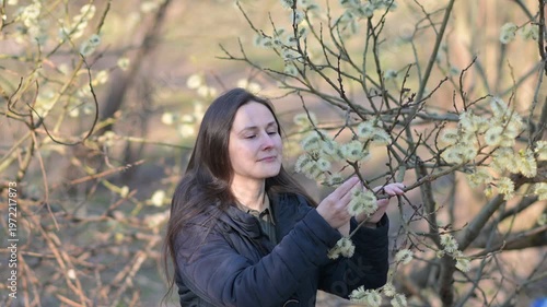 medium shot of a woman sneezing while standing close to flowering willow catkins in a spring forest. The scene clearly illustrates seasonal allergic rhinitis, hay fever symptoms