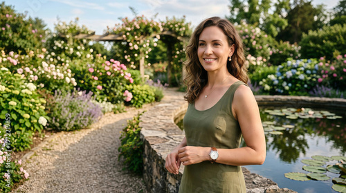 Happy woman in green dress smiling by blooming garden pond in natural sunlight